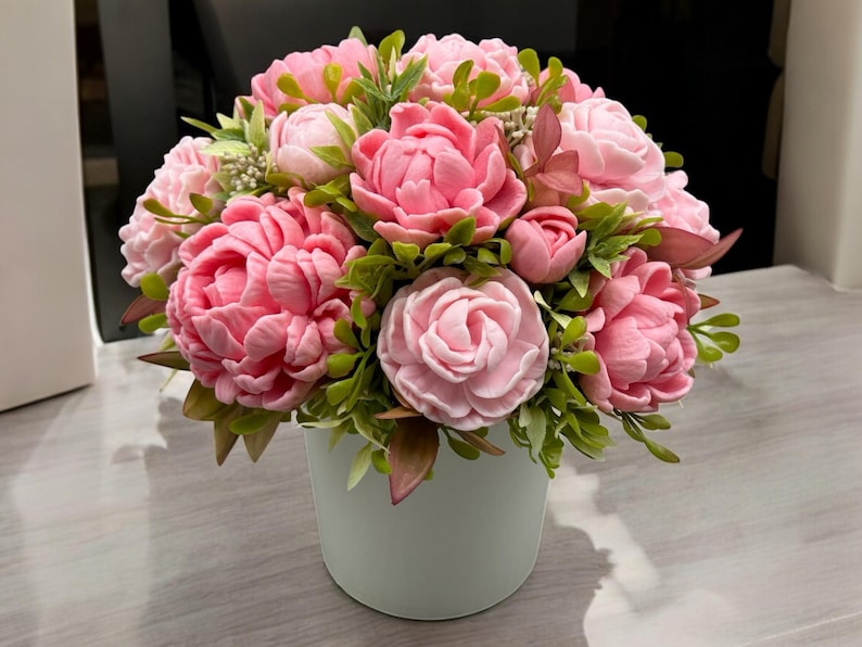 Pode incluir: Um buqu&ecirc; de flores de pe&ocirc;nia rosa em um vaso de cer&acirc;mica verde claro. As flores t&ecirc;m m&uacute;ltiplas camadas de p&eacute;talas e s&atilde;o cercadas por folhas verdes. Uma exibi&ccedil;&atilde;o floral decorativa.
