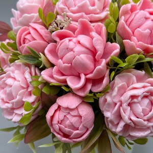Pode incluir: Um close-up de um buqu&ecirc; de pe&ocirc;nias rosa com folhas verdes e bord&ocirc;. As flores est&atilde;o em plena flora&ccedil;&atilde;o, com m&uacute;ltiplas camadas de p&eacute;talas. O arranjo est&aacute; em um vaso branco, criando uma est&eacute;tica suave e delicada.