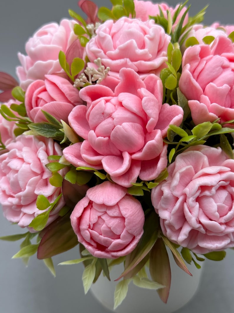 Pode incluir: Um close-up de um buqu&ecirc; de pe&ocirc;nias rosa com folhas verdes e bord&ocirc;. As flores est&atilde;o em plena flora&ccedil;&atilde;o, com m&uacute;ltiplas camadas de p&eacute;talas. O arranjo est&aacute; em um vaso branco, criando uma est&eacute;tica suave e delicada.