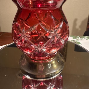 May include: A ruby red glass hurricane lamp shade featuring a deep cut starburst and diamond pattern. The glass has a wide, scalloped rim and tapers down to a circular base with a metallic finish, reflecting the intricate geometric design.