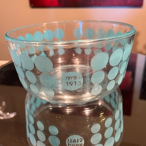 May include: Clear glass bowl with a turquoise polka dot pattern. The bowl has a rounded shape and a reflective surface. The text "Pyrex 1915" is visible on the side of the bowl. The bowl is sitting on a reflective surface.