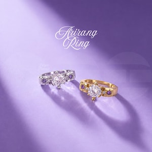 Puede incluir: Dos anillos Arirang Ring, uno de tono plateado y otro de tono dorado, cada uno con una gran piedra central transparente flanqueada por piedras moradas más pequeñas y diminutos acentos transparentes engastados en la banda sobre fondo morado.