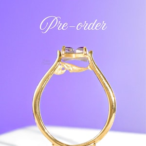 Könnte beinhalten: Ein goldener Ring mit einem quadratischen, lila Edelstein. Der Ring hat ein zartes, verziertes Design mit einem floralen Akzent am Band. Die Worte "Pre-order" sind in Kursivschrift oben im Bild geschrieben.