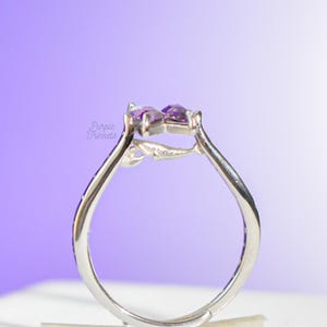 May include: A silver-toned ring featuring a small, sculpted whale figure integrated into the band, topped with two purple, heart-shaped stones. The text Purple Trinkets is visible on the left side of the ring band.