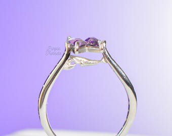 Puede incluir: Un anillo de tono plateado con una pequeña figura de ballena esculpida integrada en la banda, rematado con dos piedras moradas en forma de corazón. El texto Purple Trinkets es visible en el lado izquierdo del anillo.