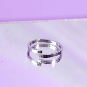 Peut inclure: Une bague argentée au design minimaliste. La bague présente un anneau fendu, créant un look ouvert et moderne. La bague est présentée sur une surface réfléchissante, avec un fond violet clair.