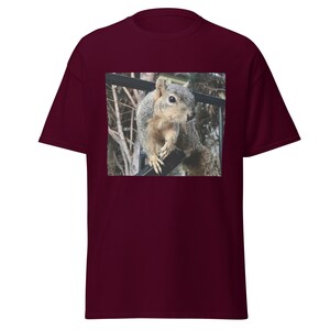 Puede incluir: Una camiseta granate con una fotografía de una ardilla posada en una barandilla negra. La ardilla tiene pelaje gris y marrón y mira hacia la izquierda. El fondo está borroso, con árboles y una valla visibles.