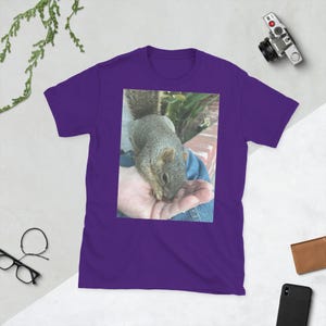 Puede incluir: Camiseta morada con una fotografía de una ardilla gris en la mano de una persona. La ardilla es el tema principal, con su pelaje y rasgos enfocados. La camiseta es de color morado liso.