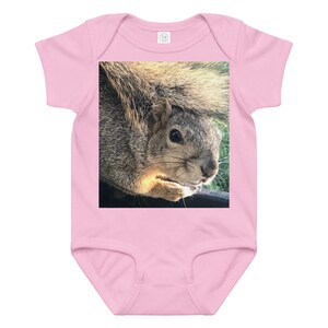 Puede incluir: Body rosa para bebé con una imagen de primer plano de una ardilla. La ardilla tiene pelaje gris y marrón, una cola esponjosa y sostiene algo en sus patas. El body está hecho de material suave.
