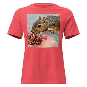 Op de afbeelding: Rood T-shirt met een vierkante afbeelding van een eekhoorn die pecannoten uit een hand eet. De eekhoorn is bruin en grijs, met een pluizige staart. De pecannoten hebben een rijke bruine kleur. Het T-shirt is gemaakt van zacht materiaal.