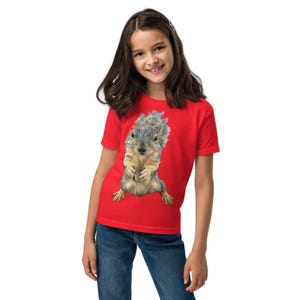 Puede incluir: Una camiseta roja con un gráfico detallado de una ardilla. La ardilla se representa en un estilo realista, con pelaje gris y marrón, de pie. La camiseta está hecha de un material suave.