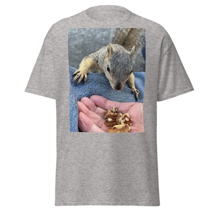 Puede incluir: Una camiseta gris con una fotografía de una ardilla mirando una mano que sostiene nueces. La ardilla tiene pelaje gris y marrón. La mano sostiene varias nueces peladas. El fondo es un par de vaqueros azules.
