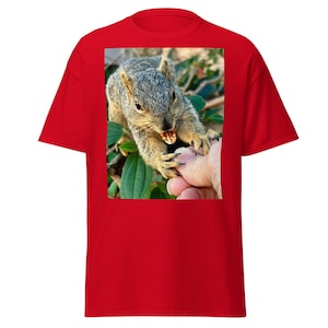 Puede incluir: Camiseta roja con una imagen de una ardilla mordiendo un dedo. La ardilla tiene pelaje gris y está rodeada de hojas verdes. La camiseta es de color rojo liso.