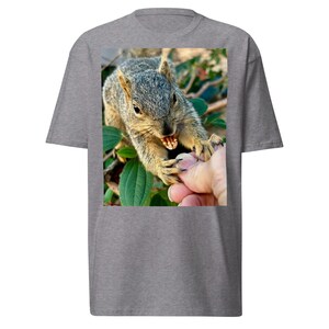 Puede incluir: Camiseta gris con una imagen de primer plano de una ardilla mordiendo el dedo de una persona. La ardilla tiene pelaje gris y está rodeada de hojas verdes. La mano de la persona es visible, con los dientes de la ardilla claramente visibles.
