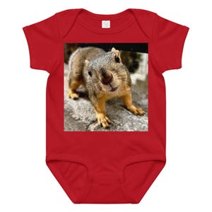Puede incluir: Body de bebé rojo con una imagen en primer plano de una ardilla. La ardilla tiene pelaje marrón y gris, con una expresión curiosa. El body está hecho de un material suave y tiene mangas cortas.