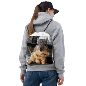 Puede incluir: Una mochila blanca con cordón con una imagen detallada de una ardilla. La ardilla está en posición sentada, con las patas juntas. La ardilla tiene pelaje gris y marrón.