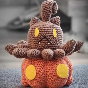 May include: A handmade crochet plush toy in the shape of a brown creature with yellow accents, resembling a cartoon character. The plush has a pumpkin-shaped base in orange with yellow circles. The toy is sitting on a gray surface.