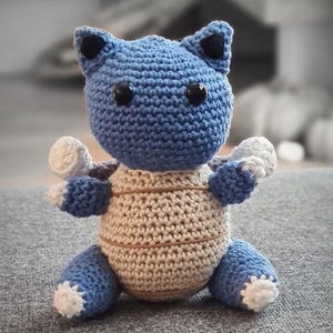 Puede incluir: Un juguete de peluche de ganchillo hecho a mano con forma de personaje de dibujos animados. El juguete presenta una cabeza y extremidades azules, un cuerpo beige y detalles blancos. Tiene ojos negros de botón y orejas pequeñas. El juguete está sobre una superficie gris.