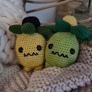 Könnte beinhalten: Zwei handgefertigte Häkelfiguren, eine gelbe und eine grüne, mit schwarzen Augen und einem Zickzack-Mund. Jede Figur hat grüne Blätter und einen schwarzen oder gelben Kopf. Sie liegen auf einer beigen Strickdecke.