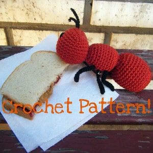 May include: A red crocheted ant with black legs is sitting on a white napkin next to a peanut butter and jelly sandwich. The text "Crochet Pattern!" is written on the napkin.