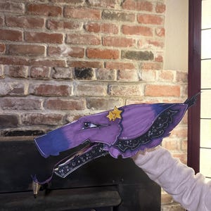 May include: A purple and blue dragon puppet featuring a stylized head with a pointed snout, a single visible eye with a dark lash, and two yellow star accents. The lower jaw is lined with a black pattern featuring white stars and swirling lines.