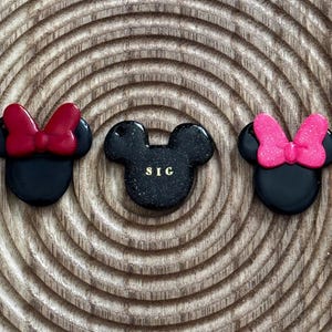 Könnte beinhalten: Drei schwarze Mickey-Mouse-Kopf-Anhänger mit roten und rosa Schleifen. Der mittlere Anhänger hat die Buchstaben "SIC" in Gold. Die Anhänger befinden sich auf einer Holzoberfläche mit kreisförmigem Maserungsmuster.