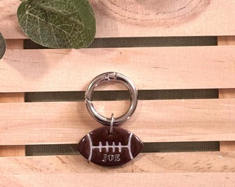 May include: A brown American football-shaped keyring with white detailing and the name "JOE" printed on it. The keyring has a silver ring clasp. The background is a wooden crate.