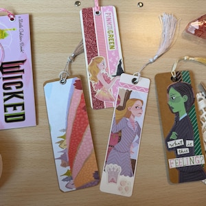 May include: A collection of decorative bookmarks with tassels. One bookmark features the word "WICKED" in large letters. Other bookmarks have illustrations of characters and the text "PINK GREEN" and "what is this FEELING?"