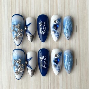 May include: A collection of ten almond-shaped false nails. Designs include blue and white ombre, silver star accents, and the text "THIS IS FOR" in white. Some nails have silver tips and jewel embellishments. The nails are arranged on a light-colored surface.