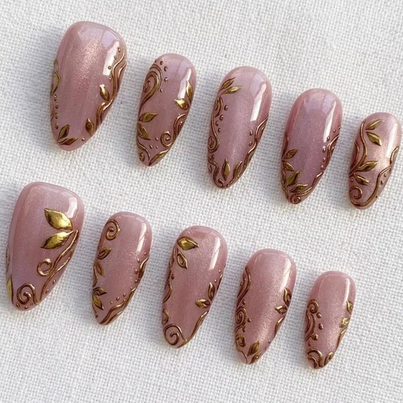 May include: Set of ten artificial nails in various sizes. Each nail is a light pink color with a gold leaf and swirl design. The nails are oval-shaped and arranged on a white surface.