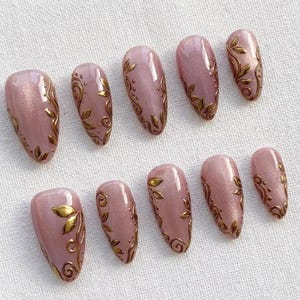 May include: Set of ten artificial nails in a soft pink hue, adorned with gold leaf and swirling patterns. The nails are oval-shaped and arranged on a white surface, showcasing a delicate and elegant design.