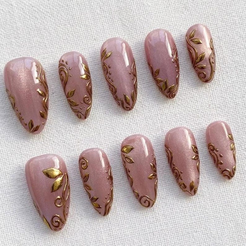 May include: Set of ten artificial nails in a soft pink hue, adorned with gold leaf and swirling patterns. The nails are oval-shaped and arranged on a white surface, showcasing a delicate and elegant design.