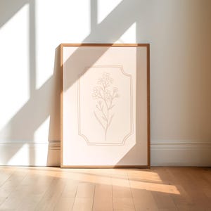 May include: A framed botanical print featuring a beige floral illustration on a cream background. The artwork is enclosed in a light brown wooden frame. The print is leaning against a white wall, with sunlight casting shadows on the wooden floor.