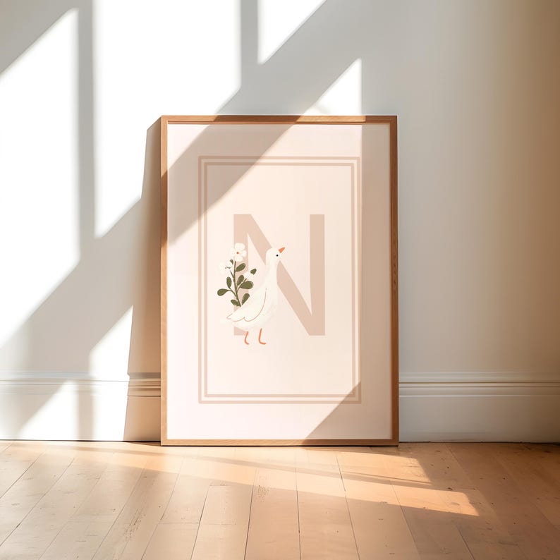 May include: A framed art print featuring a light pink background with a large letter "N" in a similar shade. A white bird with a sprig of flowers is on the left. The frame is a light brown color. The artwork is displayed on a wooden floor.