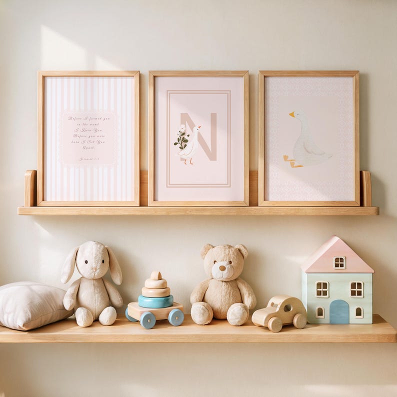 May include: A nursery shelf displays three framed prints with light wood frames. The prints feature a striped pattern, a floral letter "N", and a goose illustration. Below, a plush bunny, teddy bear, and wooden toys decorate the shelf.