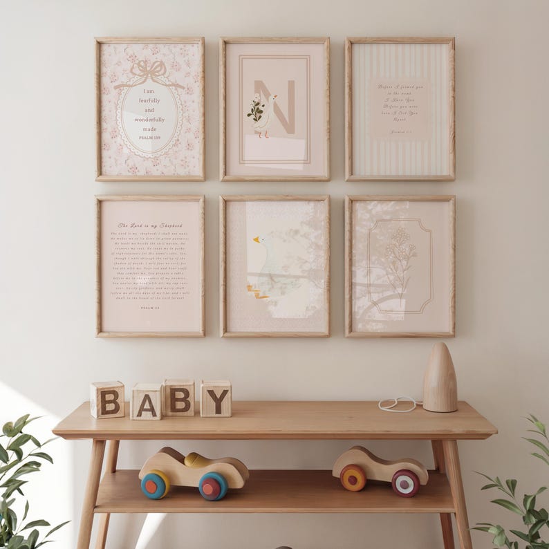 May include: A gallery wall featuring six framed prints with light wood frames. The prints include floral patterns, text, and illustrations in soft pastel colors. A wooden table below displays wooden blocks spelling "BABY" and toy cars.