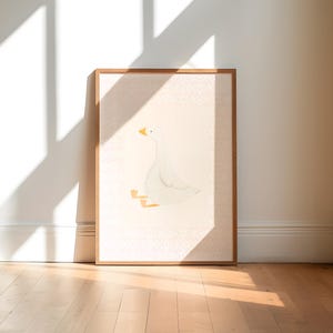 May include: A framed art print featuring a whimsical illustration of a white goose with yellow beak and feet. The goose is depicted in a seated position against a light pink background with a subtle pattern. The frame is made of wood.