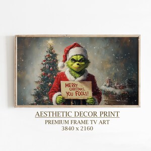 Puede incluir: Una impresión enmarcada con un personaje verde con gorro de Papá Noel y abrigo rojo, sosteniendo un letrero que dice "MERRY CHRISTMAS, YOU FOOLS!". Un árbol de Navidad y regalos están en el fondo. La impresión está etiquetada como "AESTHETIC DECOR PRINT".
