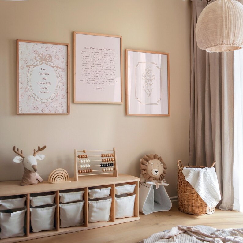 May include: A nursery scene with three framed prints on the wall. The prints have floral and text designs. Below, a wooden storage unit holds white fabric bins, a deer plush, and an abacus. A lion plush sits in a small tent.