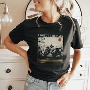 Puede incluir: Camiseta negra con el texto "PROJECT HAIL MARY" sobre un gráfico de una persona y una estructura geométrica. Debajo del gráfico están las palabras "DE GRACE & ROCKY" y rayas horizontales en naranja, amarillo y azul.