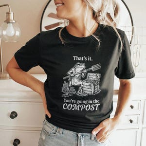 May include: Black t-shirt with a humorous graphic of a frog holding a pitchfork next to a treasure chest. The text on the shirt reads "That's it. You're going in the COMPOST."