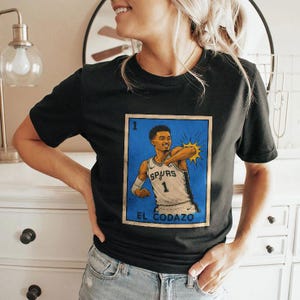 May include: Black t-shirt featuring a graphic of a basketball player in a Spurs jersey, with the number 1. The graphic is in a vintage style with the text "EL CODAZO" at the bottom. The player is depicted with a yellow burst.