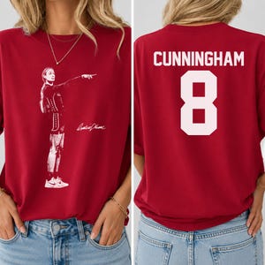 May include: A dark red t-shirt featuring a white graphic print of a basketball player pointing on the front and the name CUNNINGHAM with the number 8 in large white block lettering on the back. The shirt has a crew neckline and short sleeves.