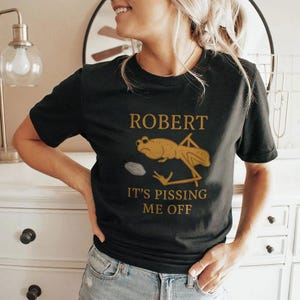 May include: Black t-shirt featuring a gold graphic of a frog and the text "ROBERT IT'S PISSING ME OFF". The frog has a grumpy expression. A casual wear item.