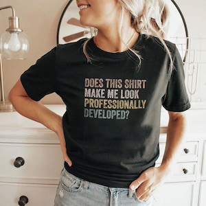 Könnte beinhalten: Ein schwarzes kurzärmeliges T-Shirt mit dem Text "DOES THIS SHIRT MAKE ME LOOK PROFESSIONALLY DEVELOPED?", der in einer gestapelten, mehrfarbigen Schrift im Vintage-Stil mit Rosa-, Gelb- und Blautönen auf dem dunklen Stoff gedruckt ist.