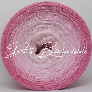 Könnte beinhalten: Eine große, runde Garnkugel mit einem Farbverlauf von Rosa und Weiß. Das Garn ist eng gewickelt und erzeugt eine glatte Oberfläche. Der Text "Danis Bobbelwerkstatt" ist in einer Schreibschrift über dem Garn geschrieben.