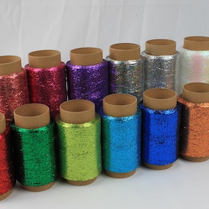 Könnte beinhalten: Eine Sammlung von vierzehn zylindrischen Spulen mit metallischem Glitzergarn in verschiedenen leuchtenden Farben wie Rot, Grün, Limette, Blau, Lila, Pink, Silber, Kupfer und Schwarz, die jeweils auf einen schlichten braunen Pappkern gewickelt sind.