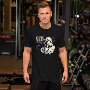 Könnte beinhalten: Ein schwarzes Kurzarm-T-Shirt mit einer weißen Grafik von Galileo Galilei, dem Text "AND YET IT MOVES EPPUR SI MUOVE ATTRIBUTED TO GALILEO GALILEI" und einer dünnen blauen Orbitallinie, die das Porträt des Wissenschaftlers umgibt.