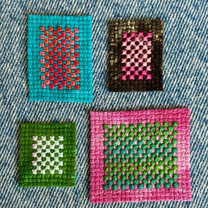 May include: Four rectangular fabric patches featuring cross-stitch embroidery. One patch is turquoise with a red and orange grid, one is black with a pink grid, one is green with a white grid, and one is pink with a green and turquoise grid.