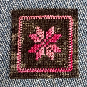 May include: A square fabric patch featuring a dark charcoal gray woven base with a pink embroidered border. The center displays a symmetrical star-like floral motif stitched in varying shades of pink thread, creating a distinct contrast against the dark background.